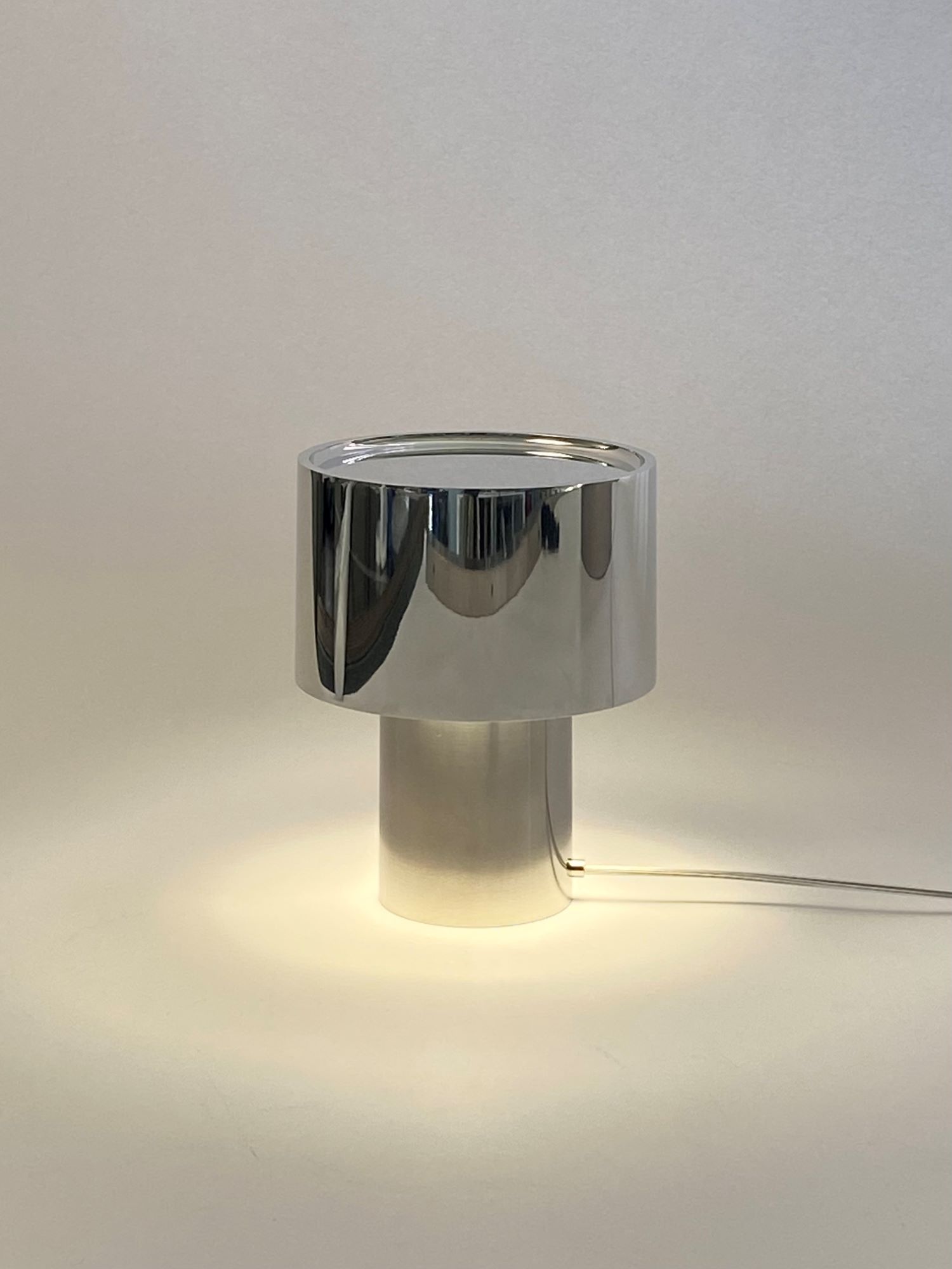 Лампа MIRROR — Настольная лампа «MIRROR» из нержавеющей стали и дюрали с цилиндрическим отражающим абажуром, светящаяся на белом фоне.
