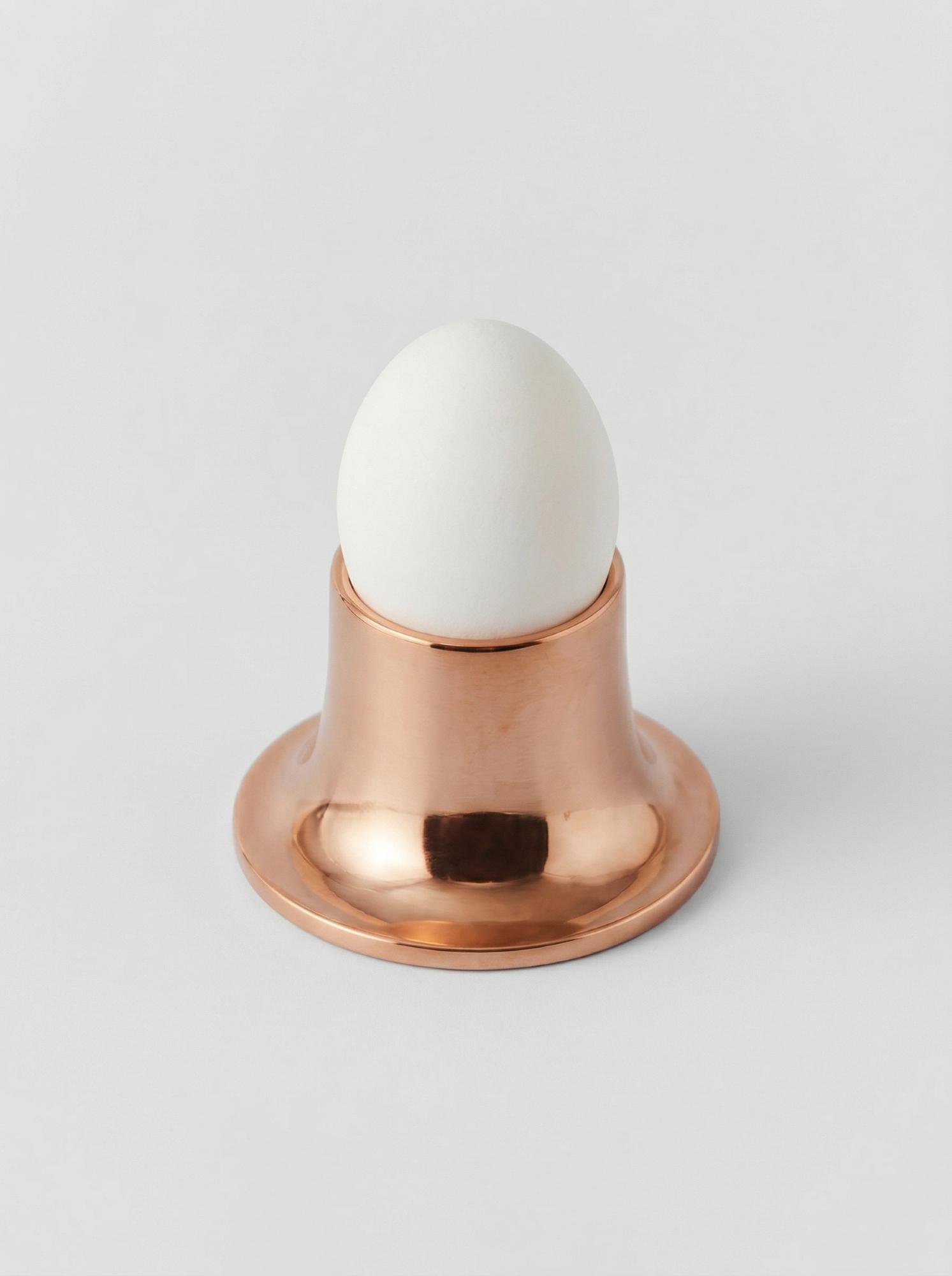 Медная подставка с гладкой полировкой, в которой установлено белое яйцо на светлом фоне